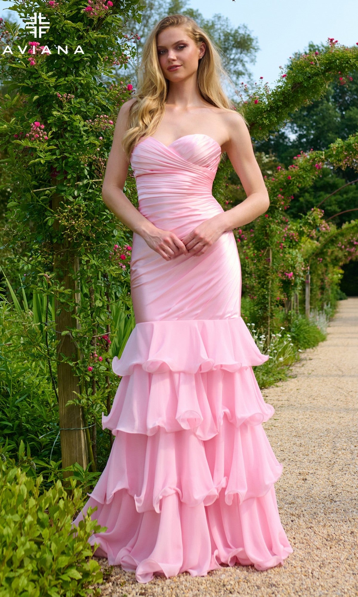 A woman stands outdoors in a light pink Faviana 11419 long prom dress featuring a strapless sweetheart neckline, ruched bodice, and layered ruffle skirt, surrounded by lush greenery and blooming flowers.